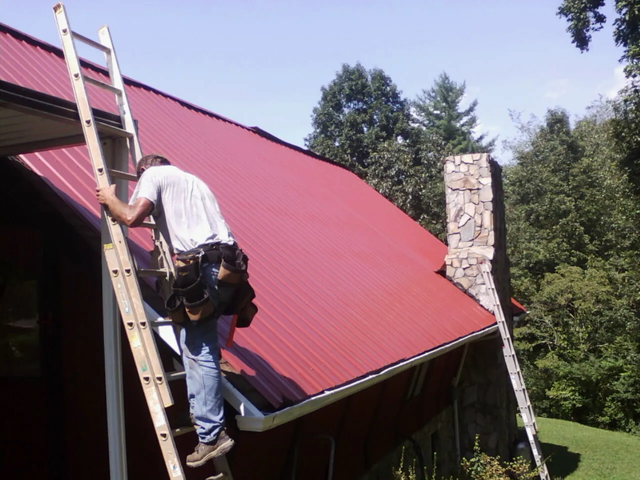 Roofer on a ladder beside a red metal roof and stone chimney — Soffit & Fascia Repair by Roofing Brothers in Portage Des Sioux